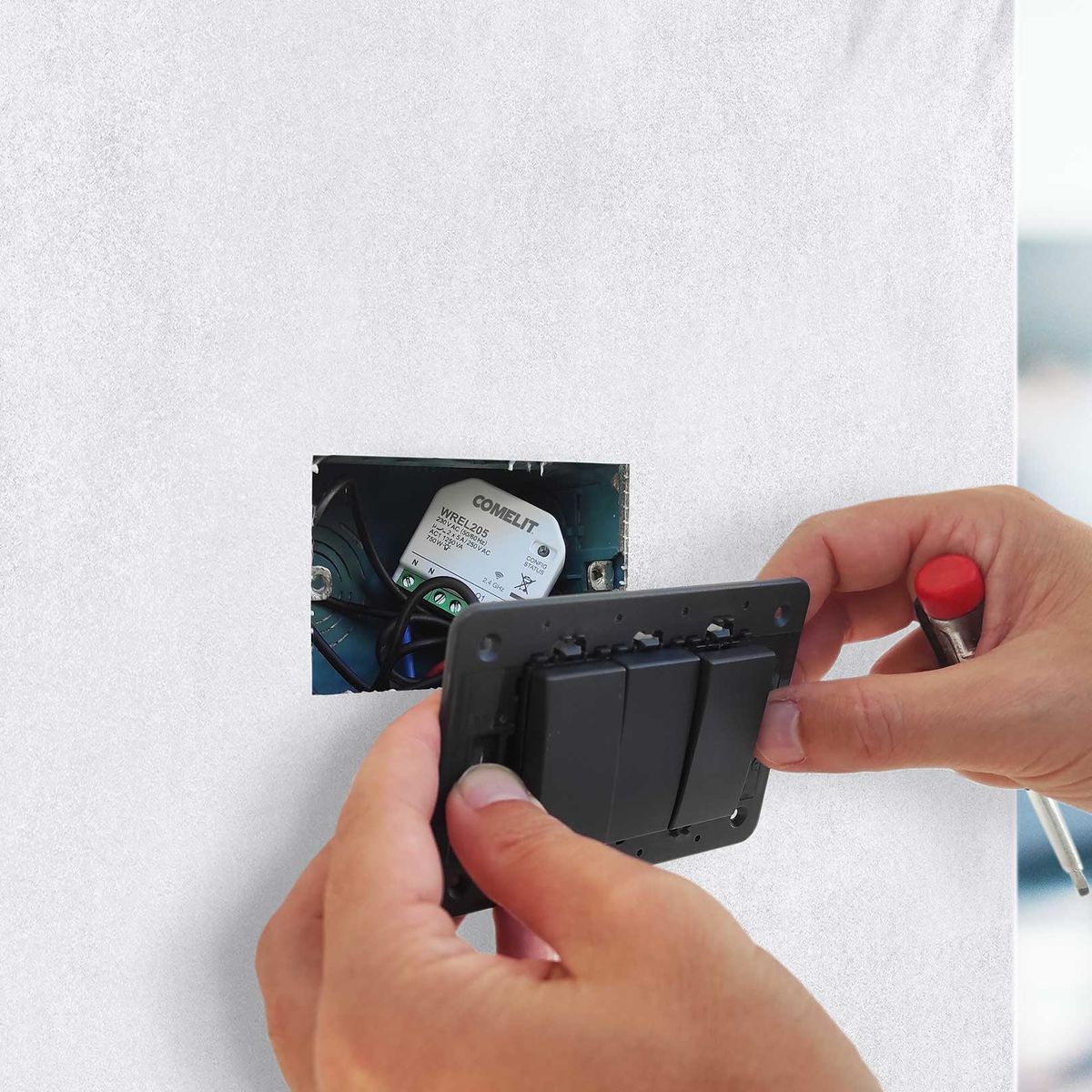Mani di un installatore che montano una placca a tre pulsanti davanti a un modulo domotico nella scatola a incasso