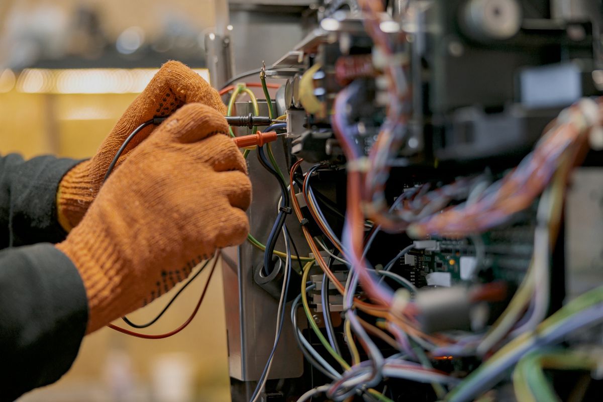 Mani con guanti da lavoro che misurano con i puntali del multimetro i collegamenti di un'apparecchiatura